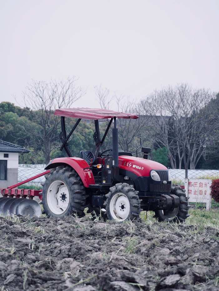 A red tractor working in a plowed field with bare trees in the background during the day.